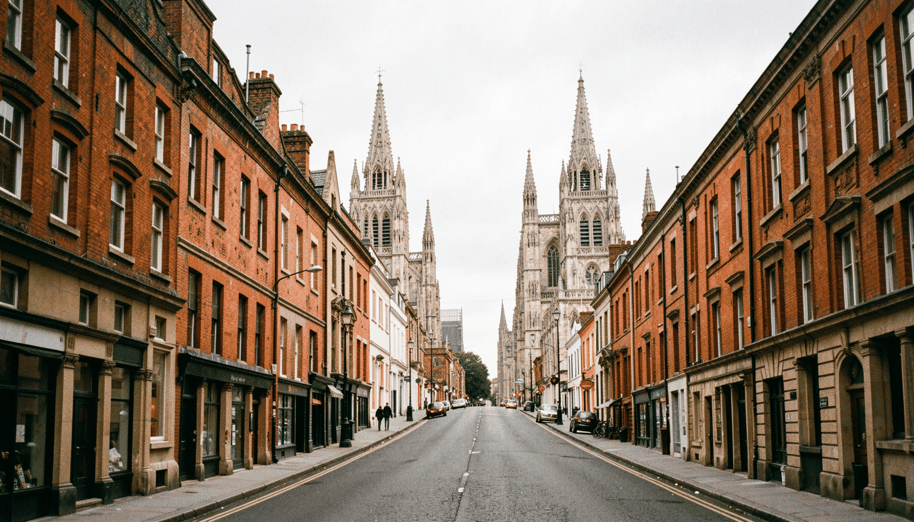 A wide street lined with historic red-brick and sandstone buildings, two grand cathedrals visible at either end under a soft grey sky